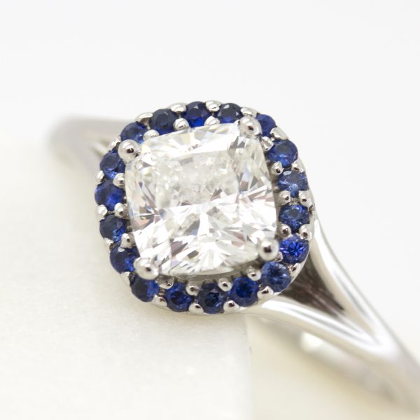 cushion cut center diamond with blue sapphire halo engagement ring set on a bias with split shank